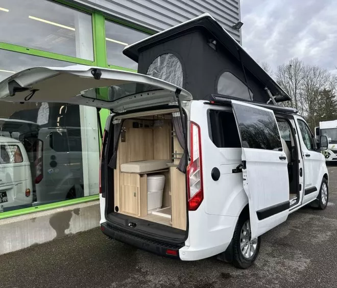 Van aménagé avec toit relevable et espace intérieur visible, stationné devant un bâtiment.