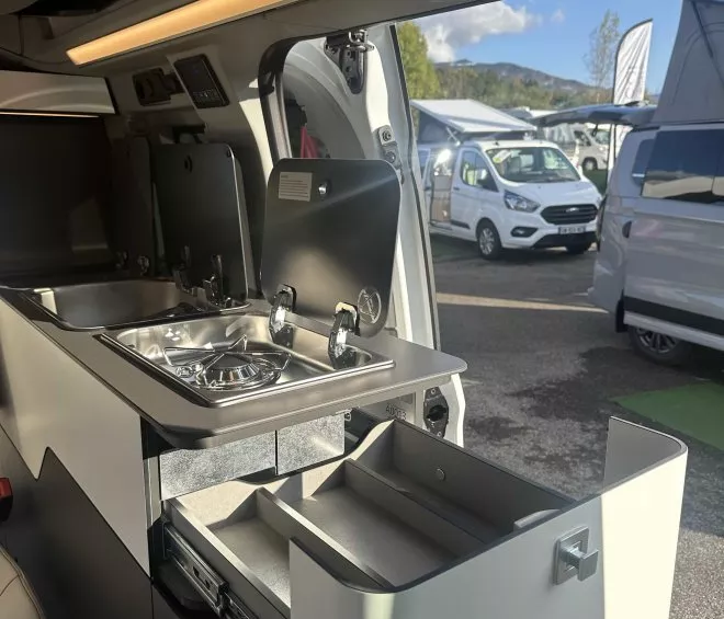 Intérieur d'un van aménagé avec évier et tiroir ouvert, camping-cars en arrière-plan