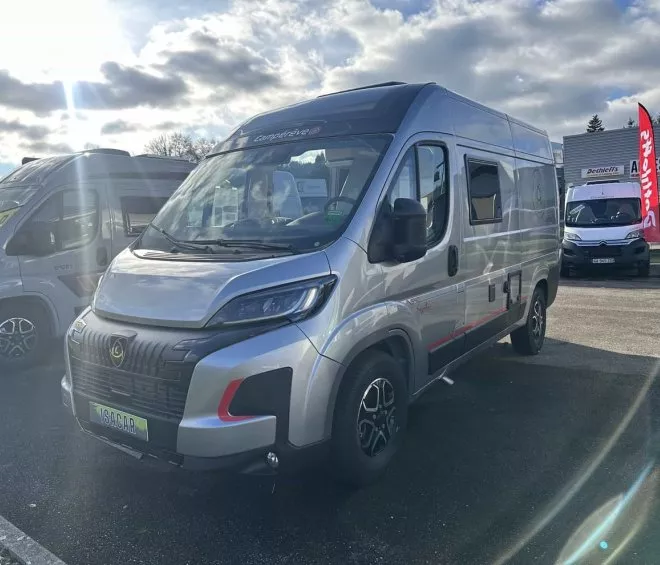 Camping-car gris stationné sur un parking ensoleillé devant un magasin d'accessoires.