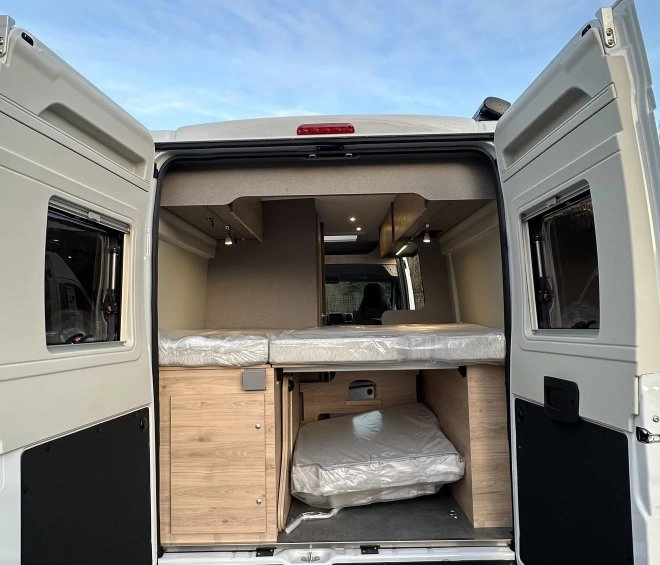 Intérieur d'un van aménagé avec lit et rangements en bois, portes arrière ouvertes