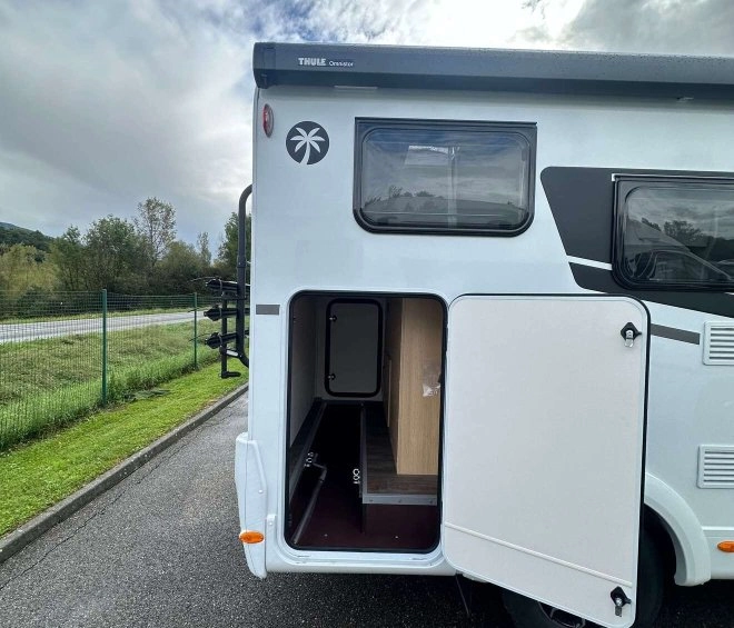 Camping-car avec porte arrière ouverte sur un parking, ciel nuageux en arrière-plan.
