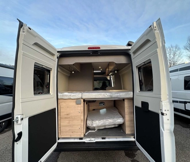 Intérieur d'un van aménagé avec portes arrière ouvertes, montrant un espace de couchage et de rangement.