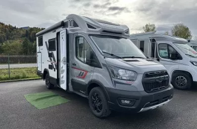 Camping-car gris stationné sur un parking, porte ouverte, ciel nuageux en arrière-plan.