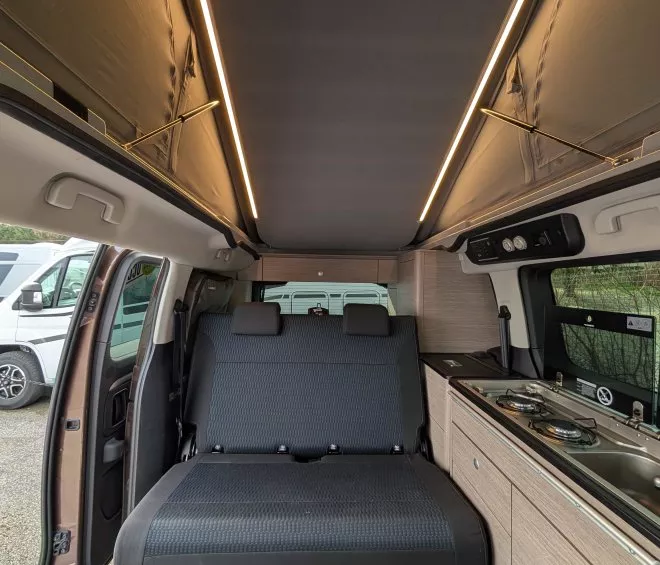 Intérieur d'un van aménagé avec banquette et cuisine intégrée, éclairage LED au plafond.