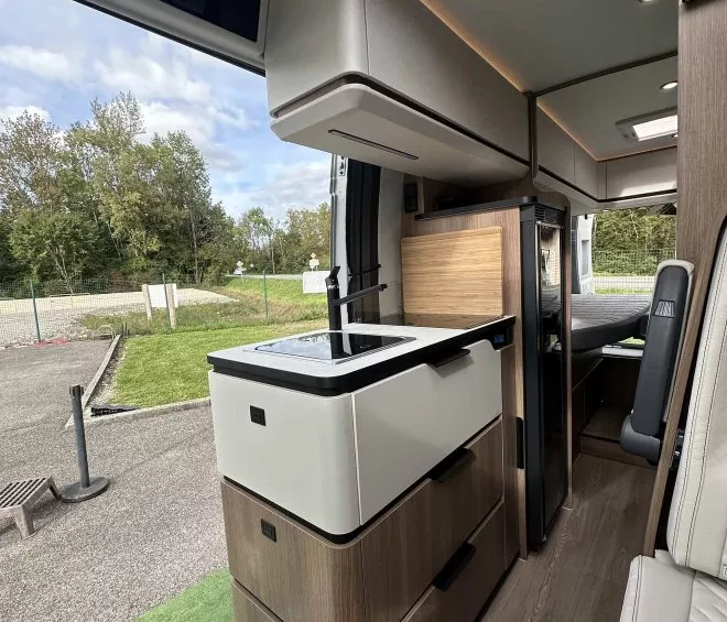 Intérieur moderne d'un camping-car avec cuisine équipée et vue sur l'extérieur verdoyant.