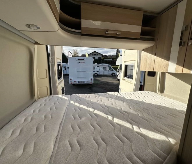 Intérieur d'un camping-car avec lit et placards en bois, vue sur d'autres véhicules stationnés.