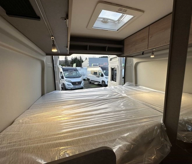 Intérieur d'un camping-car avec lit double, vue sur d'autres véhicules stationnés à l'extérieur.