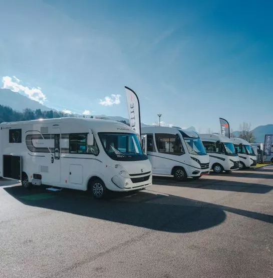 Alignement de camping-cars stationnés sur un parking sous un ciel dégagé, avec des montagnes en arrière-plan.