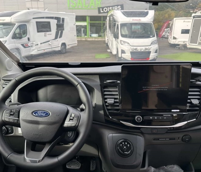 Vue intérieure d'un camping-car Ford avec écran de tableau de bord et volant, camping-cars en arrière-plan.