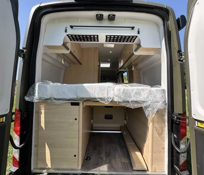 Intérieur d'un van aménagé avec lit et rangements en bois clair, portes arrière ouvertes.