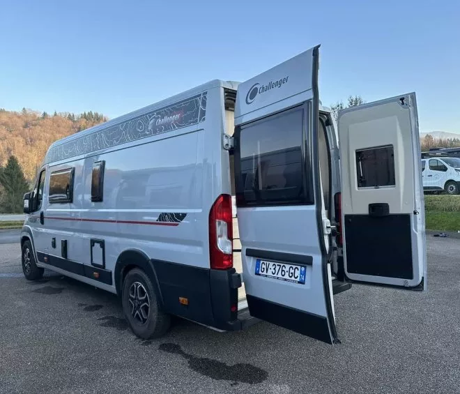 Camping-car Challenger stationné avec portes arrière ouvertes sur un parking, paysage boisé en arrière-plan.