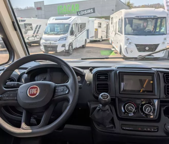 Vue intérieure d'un camping-car Fiat avec tableau de bord et volant, devant concession Isacar Loisirs.