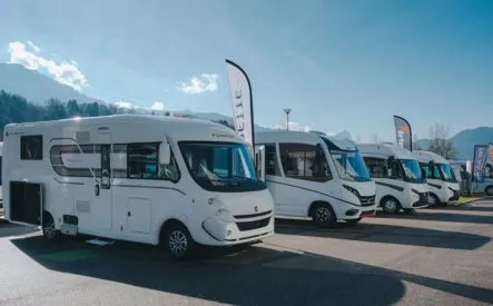 Alignement de camping-cars stationnés sur un parking sous un ciel dégagé, avec des montagnes en arrière-plan.