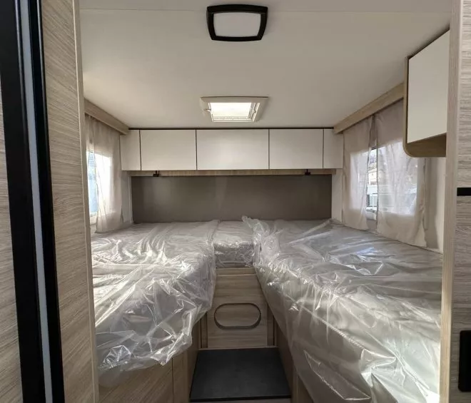 Intérieur d'une caravane avec lits jumeaux sous plastique, fenêtres latérales et placards.