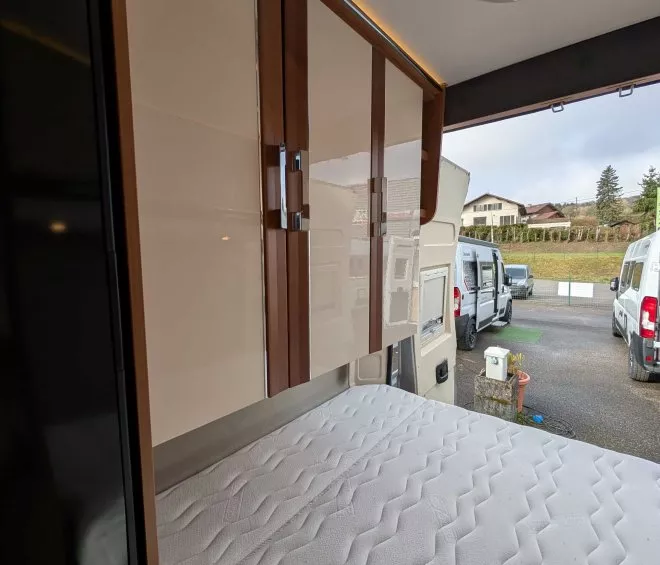 Intérieur d'un camping-car avec lit et placards en bois, stationné en extérieur.