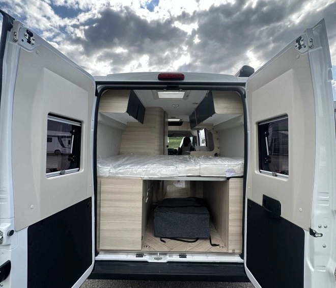 Intérieur d'un van aménagé avec portes arrière ouvertes sous un ciel nuageux.