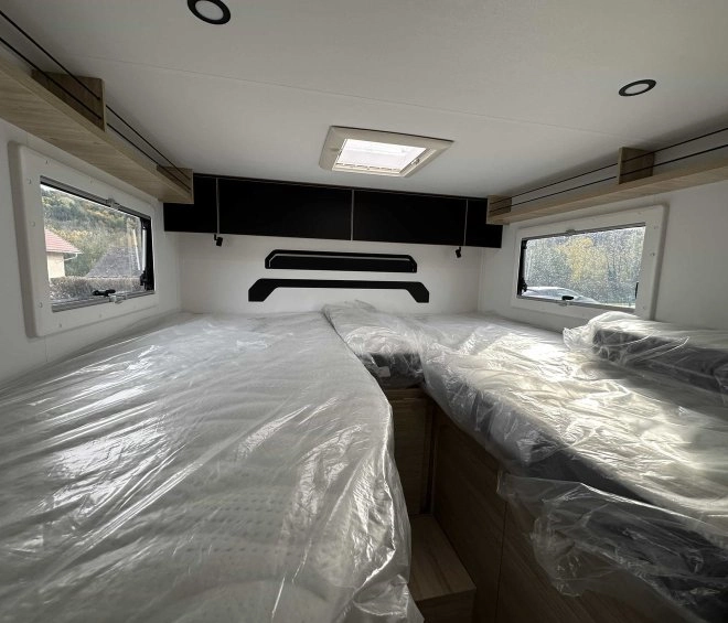 Intérieur d'un camping-car avec lits jumeaux protégés par du plastique, fenêtres latérales et rangement.