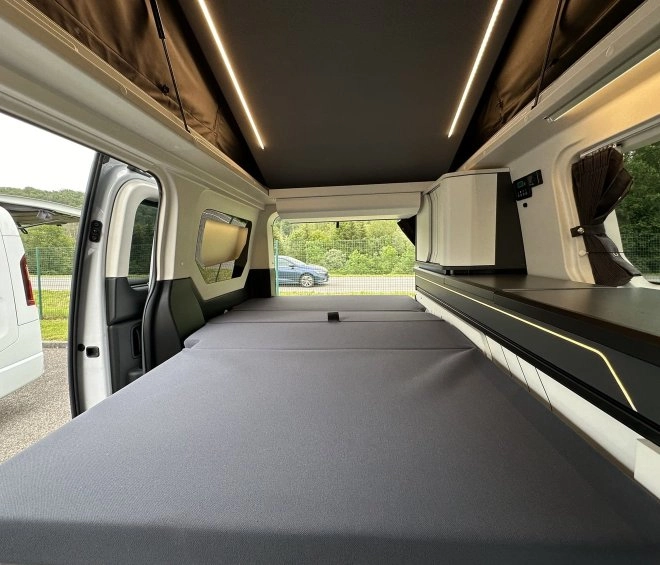 Intérieur d'un van aménagé avec lit pliable et rangements, vue sur l'extérieur