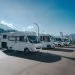 Alignement de camping-cars stationnés sur un parking sous un ciel dégagé, avec des montagnes en arrière-plan.