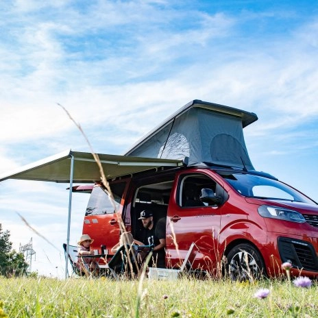 Van aménagé rouge avec toit relevable et auvent, personnes assises à l'extérieur