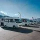Alignement de camping-cars stationnés sur un parking sous un ciel dégagé, avec des montagnes en arrière-plan.