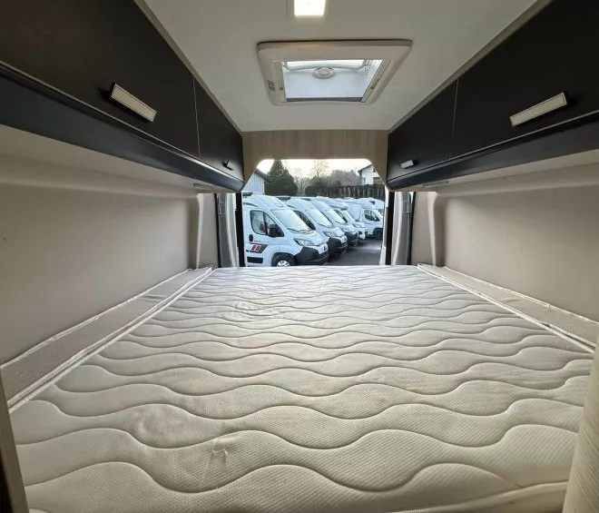 Intérieur d'un camping-car avec lit et rangements, vue sur un parking de véhicules.