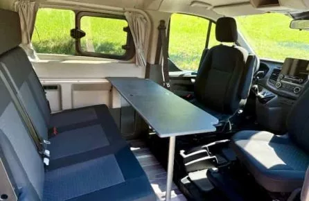 Intérieur d'un van aménagé avec sièges et table pliante, vue sur l'extérieur verdoyant.