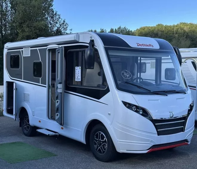 Camping-car Dethleffs blanc stationné sur un parking, porte ouverte, ciel bleu en arrière-plan.