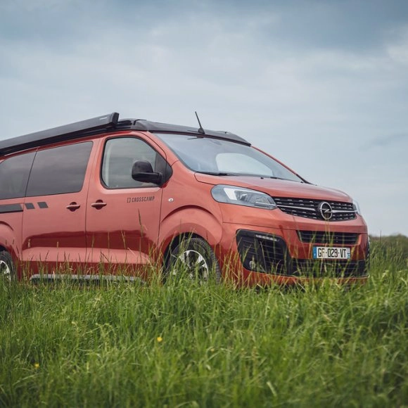 Van Crosscamp rouge stationné dans un champ verdoyant sous un ciel nuageux.