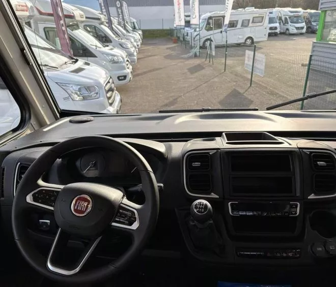 Vue intérieure d'un camping-car Fiat avec tableau de bord et volant, alignement de véhicules en arrière-plan.