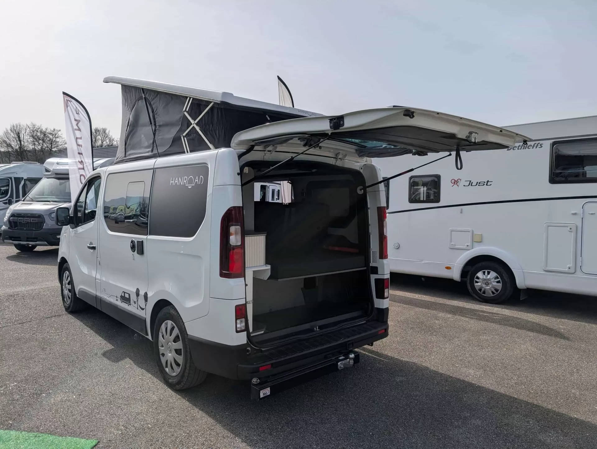 Véhicule aménagé Hanroad avec toit relevable et hayon ouvert sur un parking de camping-cars.