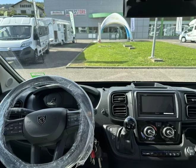 Vue intérieure d'un camping-car Peugeot avec tableau de bord et volant sous plastique, stationné devant un magasin.