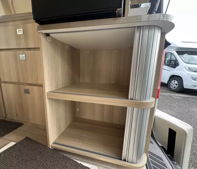 Étagère en bois clair dans un camping-car, avec rangement intégré et véhicule visible à l'extérieur.