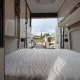 Intérieur d'un camping-car avec lit et rangements, porte arrière ouverte sur un parking.