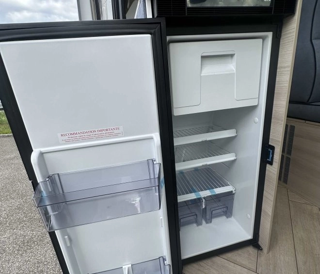 Réfrigérateur ouvert dans un camping-car, montrant des étagères vides et un intérieur spacieux.