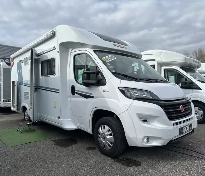 Camping-car blanc stationné sur un parking, modèle Pilote, ciel nuageux en arrière-plan.