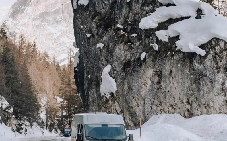 Fourgon gris sur route enneigée en montagne, entouré de sapins et de rochers