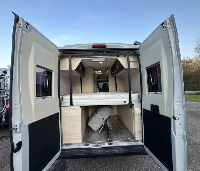 Intérieur d'un van aménagé avec portes arrière ouvertes, montrant espace de rangement et lit.