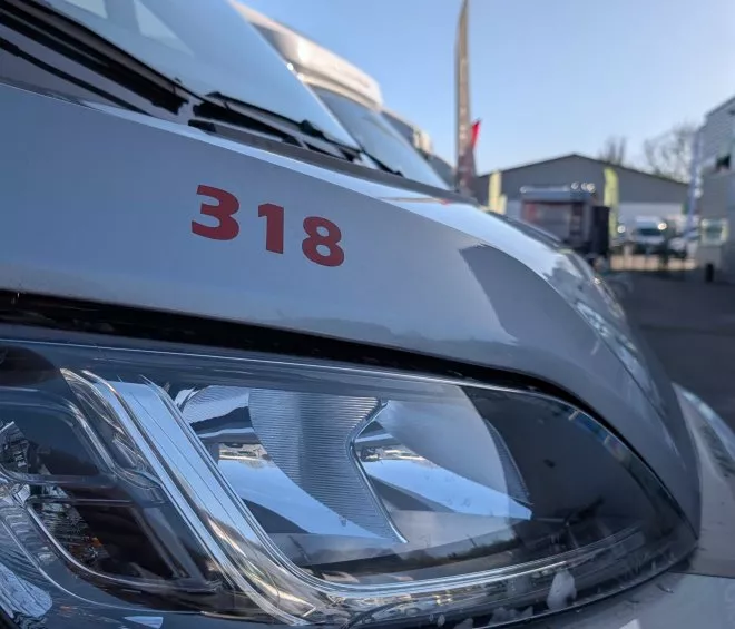 Gros plan sur le phare avant d'un véhicule utilitaire gris avec le numéro 318, stationné devant un bâtiment.