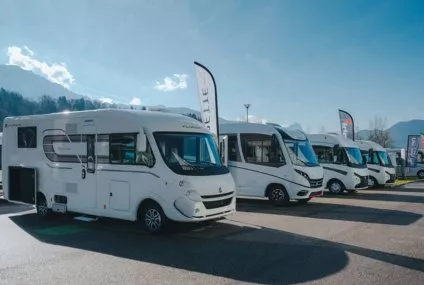 Alignement de camping-cars stationnés sur un parking sous un ciel dégagé, avec des montagnes en arrière-plan.