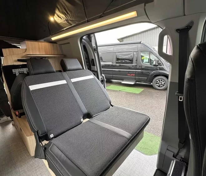 Intérieur d'un van aménagé avec sièges rabattables et vue sur un autre van à l'extérieur.