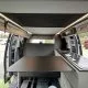 Intérieur d'un van aménagé avec table et cuisine intégrée, portes ouvertes sur un parking