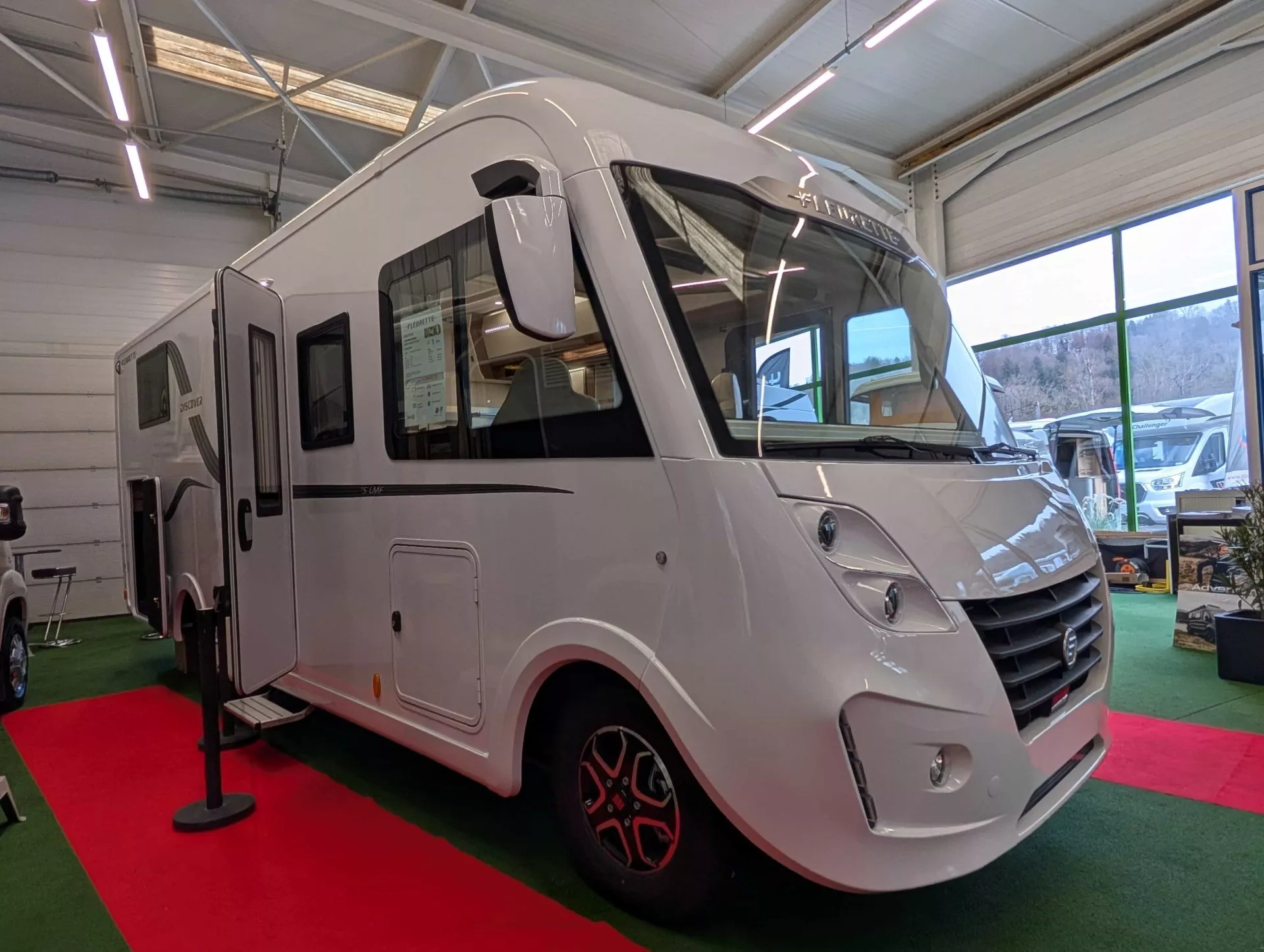 Camping-car blanc exposé dans un showroom lumineux avec sol rouge et vert.