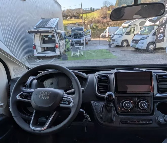 Vue intérieure d'un camping-car Fiat avec tableau de bord et véhicules stationnés à l'extérieur