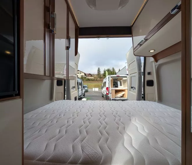 Intérieur d'un camping-car avec lit et rangements, porte arrière ouverte sur un parking.