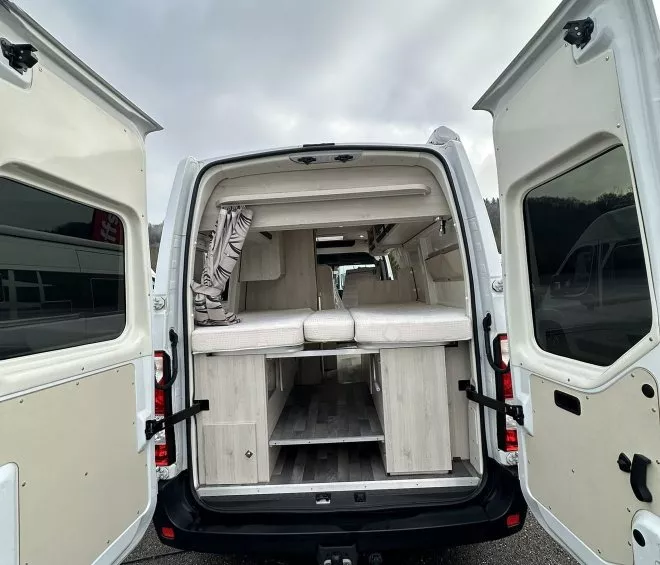 Intérieur aménagé d'un van avec portes arrière ouvertes, montrant des rangements et un espace de couchage.