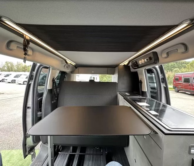 Intérieur d'un van aménagé avec table et cuisine intégrée, portes ouvertes sur un parking