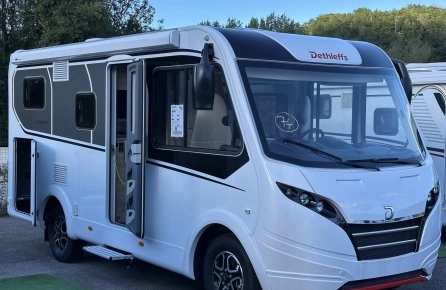 Camping-car Dethleffs blanc stationné sur un parking, porte ouverte, ciel bleu en arrière-plan.