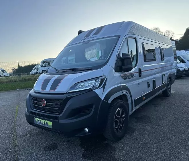 Camping-car gris stationné sur un parking avec d'autres véhicules similaires en arrière-plan.
