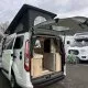 Van aménagé avec toit relevable et intérieur visible, stationné sur un parking avec d'autres camping-cars.